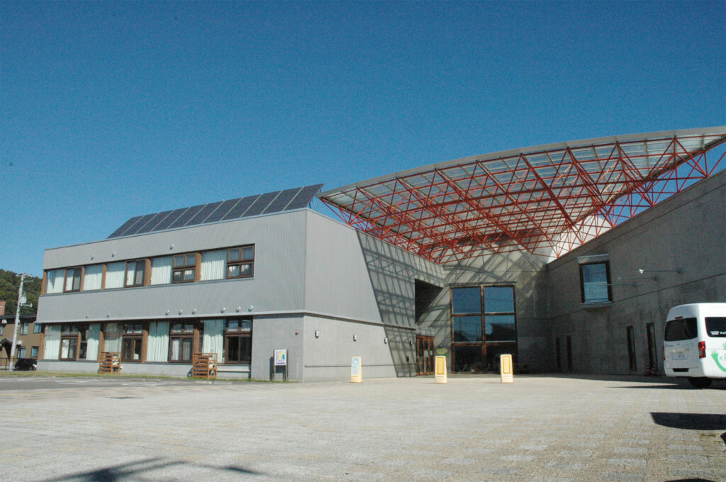 介護施設の建物外観。モダンな白壁と赤色鉄骨の大型増築棟が特徴。